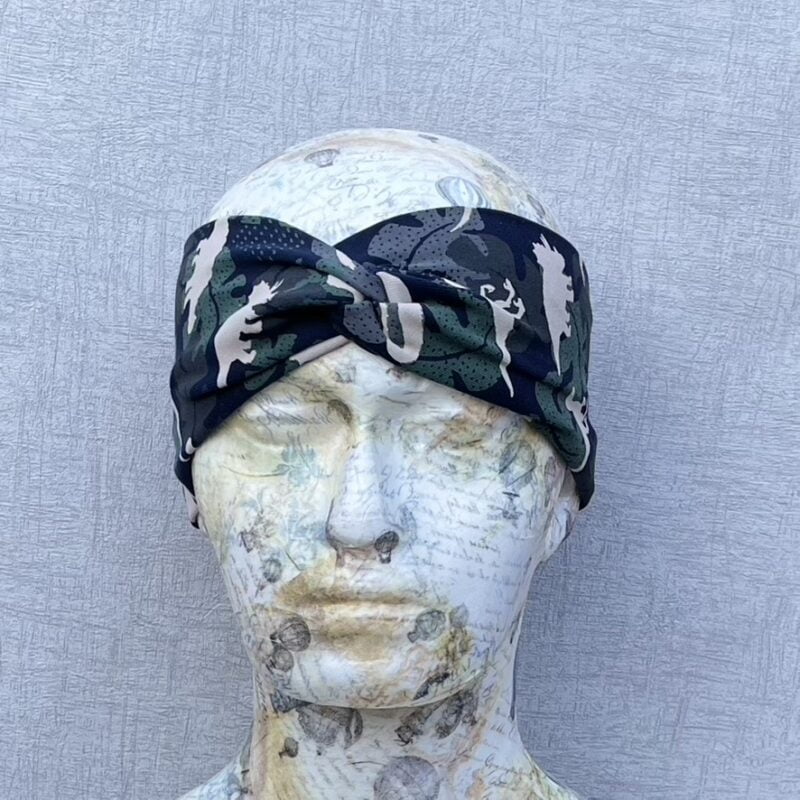 Jersey Twisted Headband in green, grey and brown camouflage colours with grey dinosaurs, randomly placed. Include Triceratops, stegosaurus, T-rex and pterodactyl. Placed on a polystyrene head that has been decoupaged with pale white and yellow paper depicting various instruments, music and corsetry. Grey background.