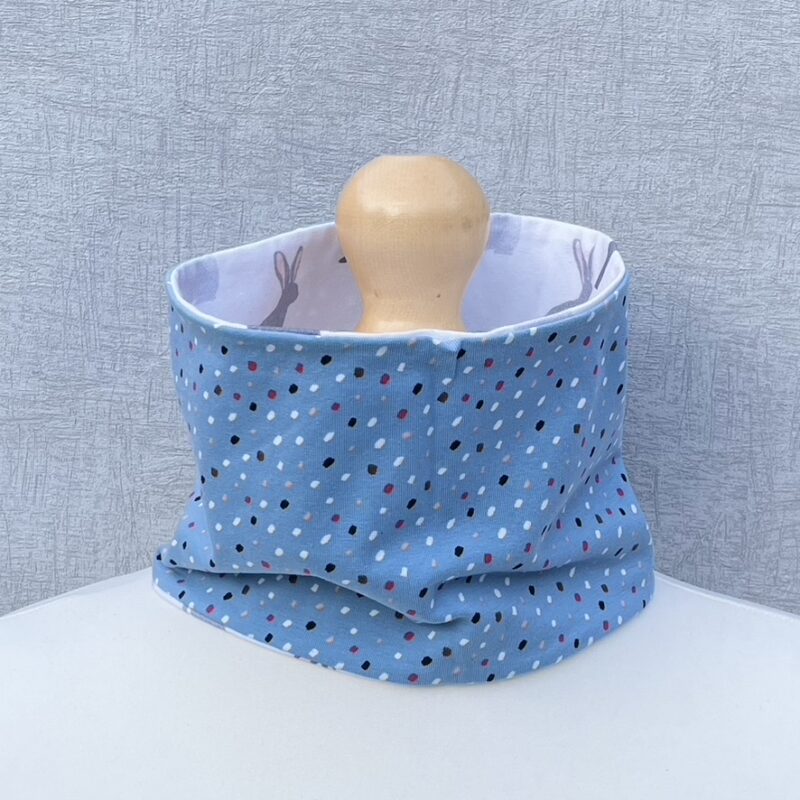 Jersey Neck Gaiter with closely spaced Polka dots in white, red, black and navy blue on a blue background. On a mannequin with pale shoulders and a wooden topper. Light grey background.