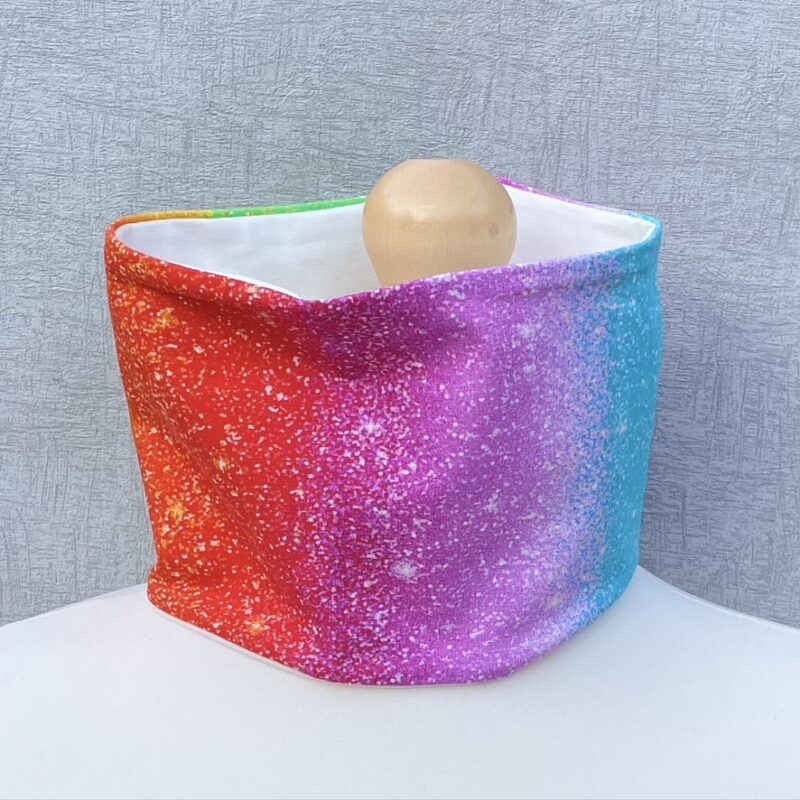 Jersey Neck Gaiter with Rainbow with glitter style dots design jersey with red, pink, blue, orange, yellow and green wide vertical stripes. On a mannequin with pale shoulders and a wooden topper. Light grey background.