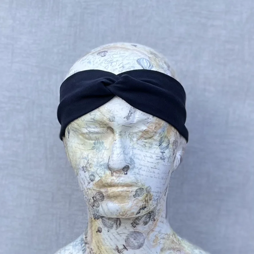 Jersey Twisted Headband in plain black. Slightly slimmer than some. Placed on a polystyrene head that has been decoupaged with pale white and yellow paper depicting various instruments, music and corsetry. Grey background.