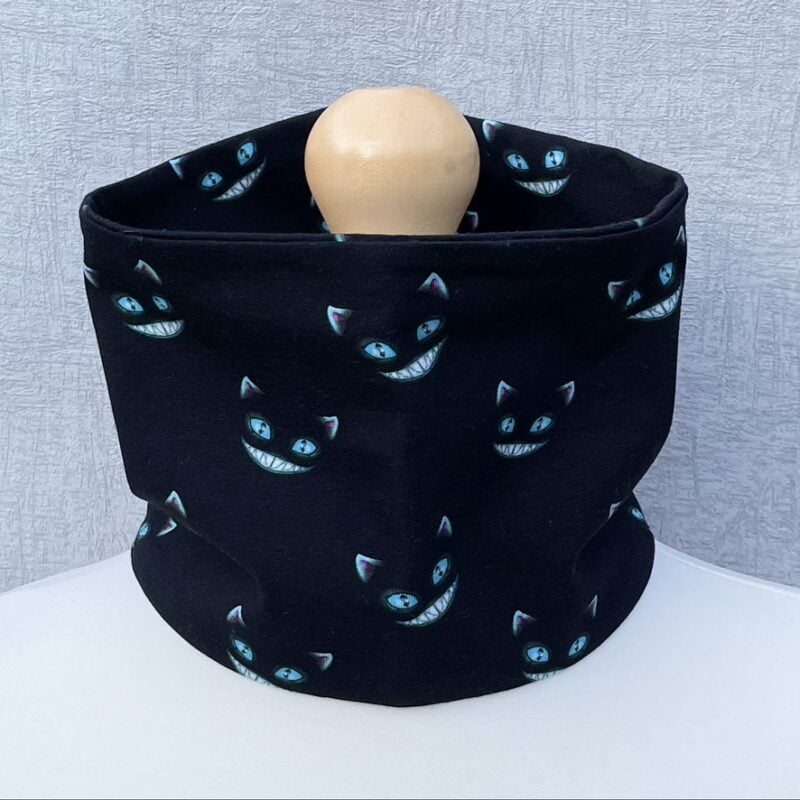 Jersey Neck Gaiter with grinning ghost cats showing white teeth, blue eyes and light blue edged ears with a pink inner ear. Black background. On a mannequin with pale shoulders and a wooden topper. Light grey background