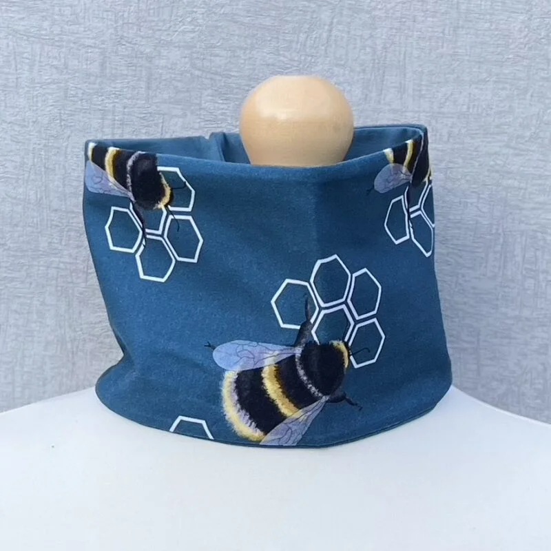 Neck Gaiter in double jersey material with a design of bees and honeycombs. Bees are sitting on the honeycomb. Background is blue. Bees are bubble bees with detailed wings. On a mannequin with pale shoulders and a wooden topper. Light grey background.