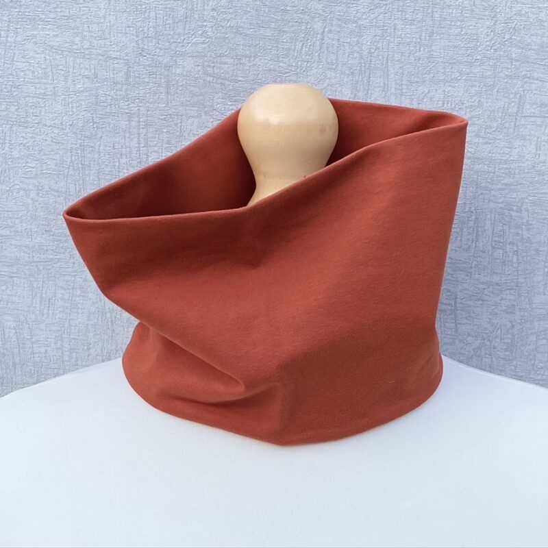 Terracotta colour neck gaiter, double jersey, sat on a mannequin with pale shoulders and a wooden topper.