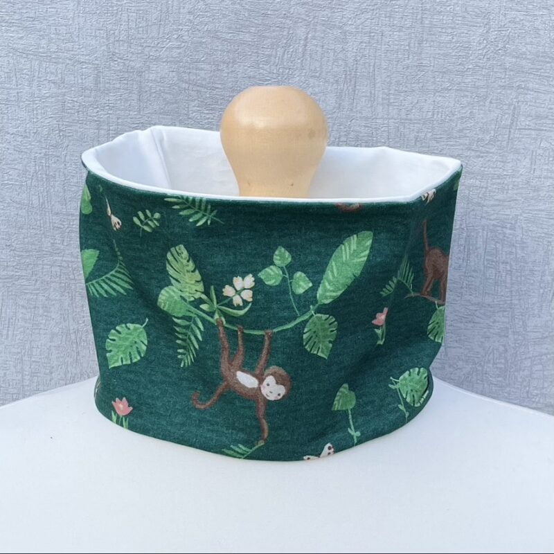 Jersey Neck Gaiter with jungle leaves, light green ferns, brown monkeys with pale faces and chests and brown feathered parrots. Pale flowers and butterflies randomly placed. Monkeys are climbing and hanging. Green background. On a mannequin with pale shoulders and a wooden topper. Light grey background.